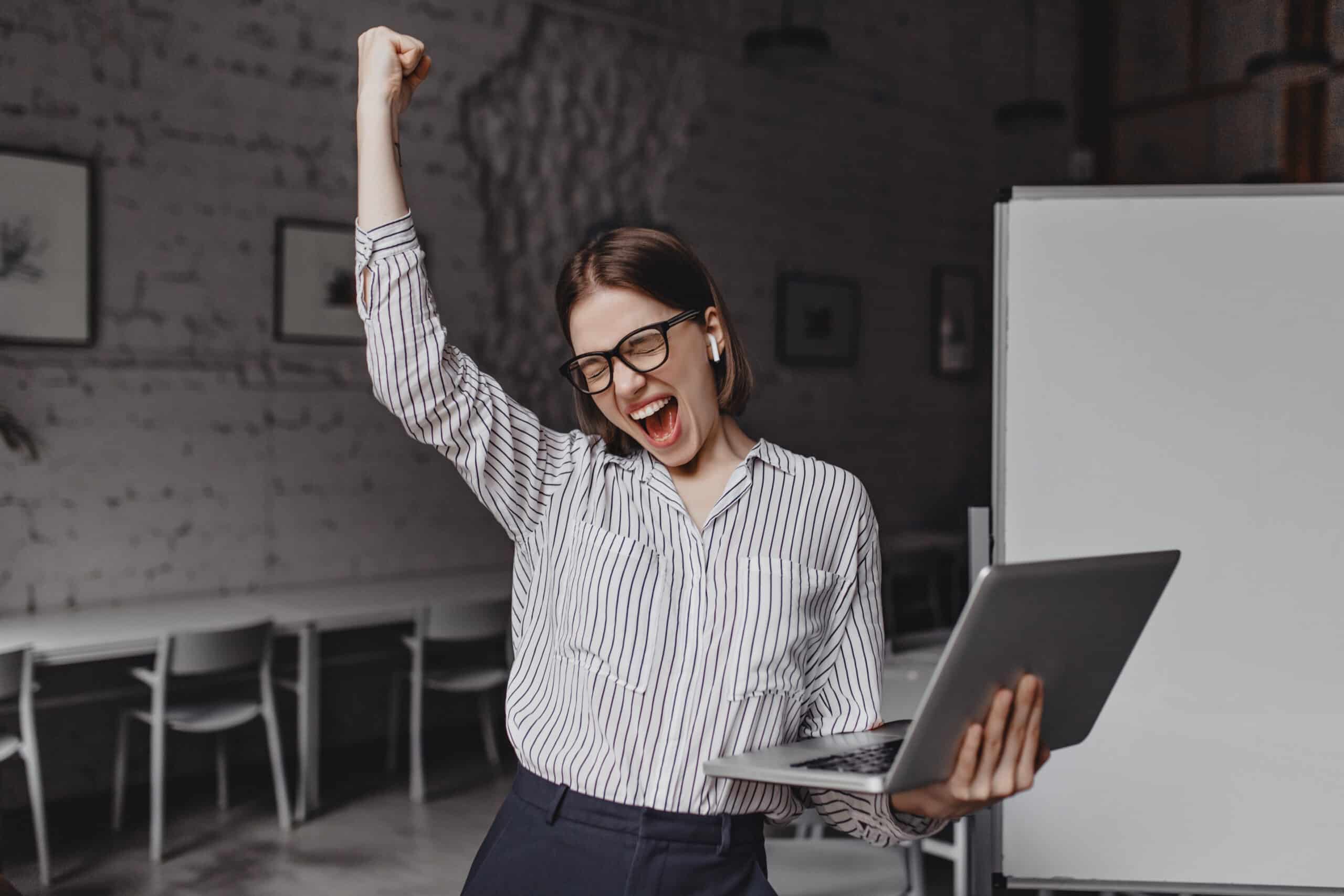 Business woman with laptop in hand is happy with success Portrait of woman in glasses and striped blouse enthusiastically screaming and making winning gesture